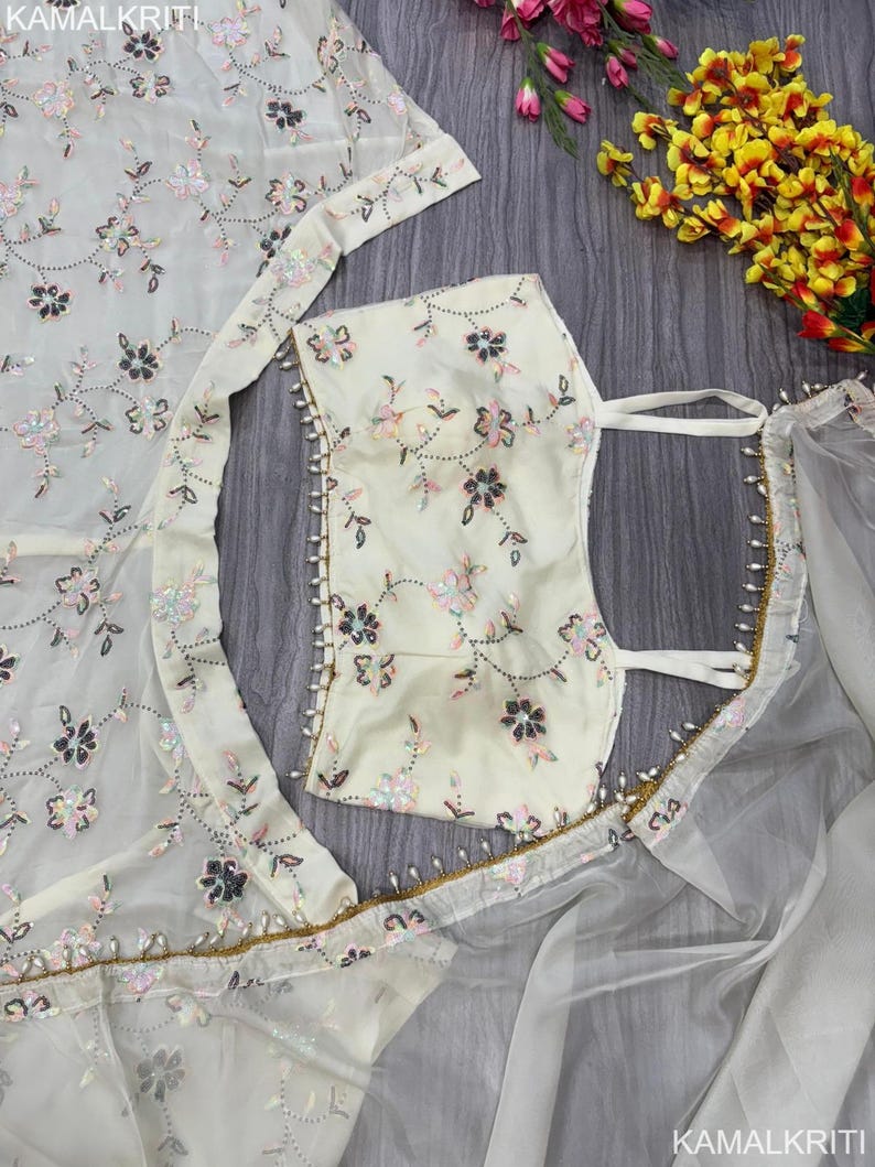 May include: Ivory-colored embroidered fabric with floral patterns. The fabric is adorned with sequins and beads, and is part of a garment. The image also shows a matching top and a sheer overlay, all with delicate floral embroidery.
