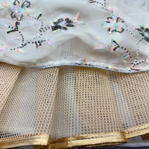 May include: Close-up of a cream-colored fabric with floral embroidery and sequin embellishments. The design features a layer of sheer fabric over a gold-toned mesh, with a gold trim at the bottom. The fabric is likely part of a dress or garment.