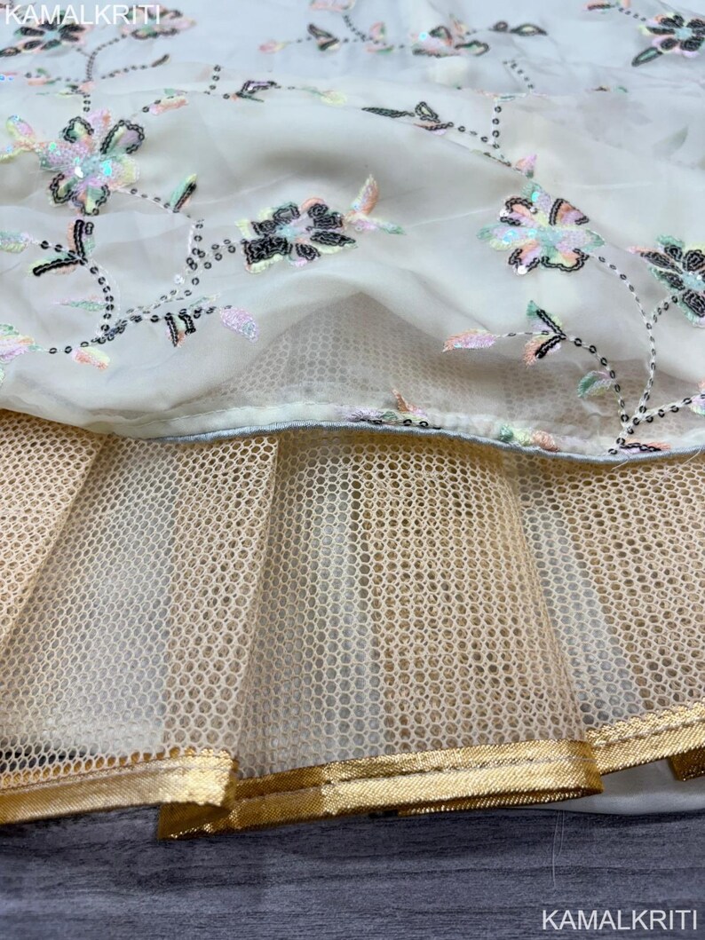 May include: Close-up of a cream-colored fabric with floral embroidery and sequin embellishments. The design features a layer of sheer fabric over a gold-toned mesh, with a gold trim at the bottom. The fabric is likely part of a dress or garment.