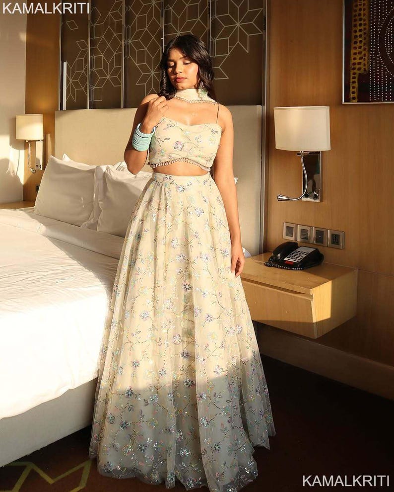 May include: A cream-colored two-piece outfit with floral embroidery. The top is a crop top with spaghetti straps and a matching choker. The skirt is a long, flowing skirt. The outfit is worn by a person in a hotel room. The text "KAMALKRITI" is visible.