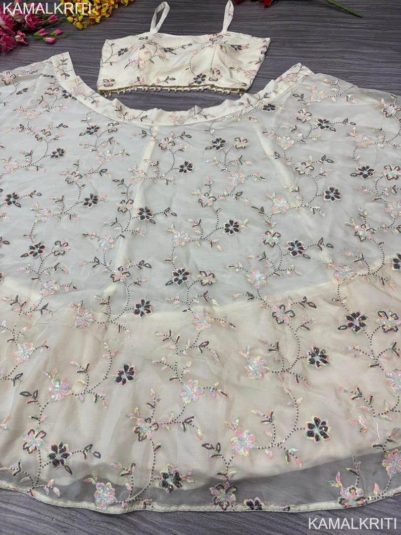 May include: Ivory-colored formal wear set featuring a crop top and a flowing skirt. The fabric is adorned with delicate floral embroidery in shades of pink, black, and silver sequins. The top has thin straps and a beaded trim.