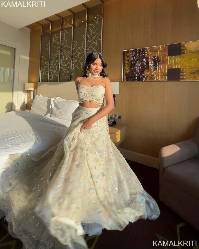 May include: A cream-colored, two-piece formal gown with delicate floral embroidery and sparkling accents. The top is a crop top with spaghetti straps, and the skirt flows to the floor. The image includes the text "KAMALKRITI".