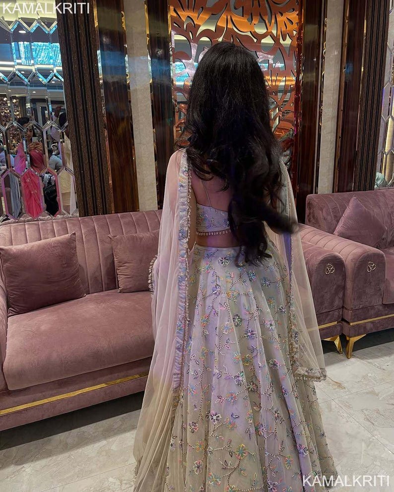 May include: A light grey lehenga with colorful floral embroidery. The outfit includes a matching top and a sheer dupatta. The text "KAMALKRITI" is visible in the bottom right corner.