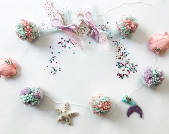 Könnte beinhalten: Eine verspielte Girlande mit Pompons in Rosa, Mintgrün und Lavendel, durchsetzt mit Filz-Meerjungfrauenschwänzen, Seesternen und Muscheln. Die Girlande ist mit Glitzer und Bändern verziert und schafft ein festliches Dekorationselement.