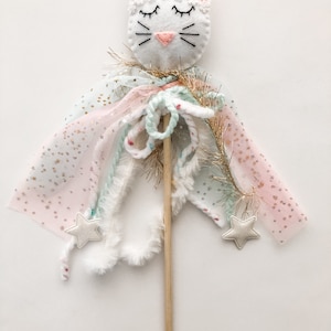 May include: A white felt cat head with pink ears and closed eyes is attached to a wooden stick. The stick is decorated with pink, white, and green tulle, white fur, and gold glitter. Two white felt stars are attached to the tulle.