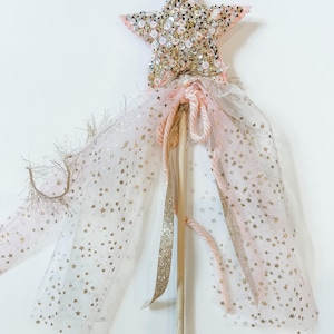 May include: A whimsical fairy wand with a pink star-shaped top adorned with sequins. The wand features a wooden dowel, pink and gold accents, and a flowing tulle skirt sprinkled with gold stars. A charming accessory for imaginative play.