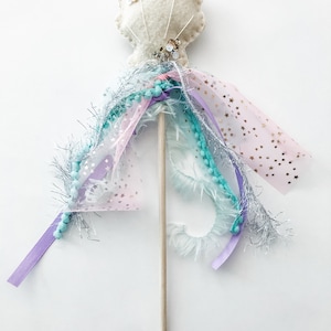 May include: A white felt seashell with gold glitter and sequins attached to a wooden stick. The stick is decorated with pink, purple, and blue ribbons, tulle, and beads.