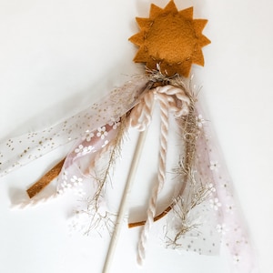May include: A whimsical wand with a star-shaped felt topper in a mustard yellow color. The wand is adorned with pink and white tulle, gold glitter ribbon, and a beige rope.