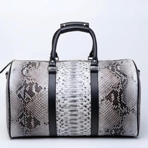 May include: A duffel bag with a snakeskin pattern in shades of gray and white. The bag has black leather handles and straps. The zipper is black. The bag is designed for travel and has a cylindrical shape.