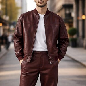 May include: A burgundy leather bomber jacket and matching pants are worn with a white t-shirt and white sneakers. The jacket has a zip closure and ribbed cuffs. The pants are straight-legged.