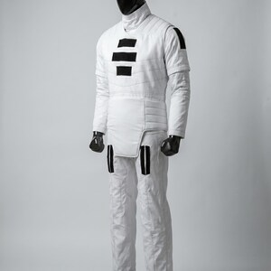 May include: A white quilted jumpsuit with black accents, displayed on a mannequin. The suit features a high collar, horizontal black stripes on the chest, and vertical black stripes on the legs. The mannequin has a black head and hands.