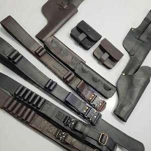 K&ouml;nnte beinhalten: Eine Auswahl handgefertigter Lederholster, G&uuml;rtel und Taschen in verschiedenen Braun- und Schwarzt&ouml;nen. Das Bild zeigt verschiedene Designs, darunter Waffenholster, Patroneng&uuml;rtel und kleine Taschen mit Schnallen- und Riemenverschl&uuml;ssen.