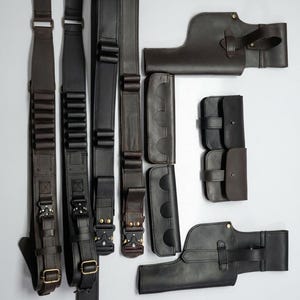 K&ouml;nnte beinhalten: Eine Auswahl an dunkelbraunen und schwarzen Leder-Waffenzubeh&ouml;r, darunter Riemen, Patronenhalter und Holster. Die Artikel sind auf einer wei&szlig;en Oberfl&auml;che angeordnet und zeigen die verschiedenen Designs und Hardware-Details.