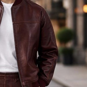 May include: A burgundy leather bomber jacket with a zip closure and ribbed cuffs. The jacket is worn over a white t-shirt. The jacket's color is a deep, rich shade of burgundy, and the leather appears to have a slight sheen.