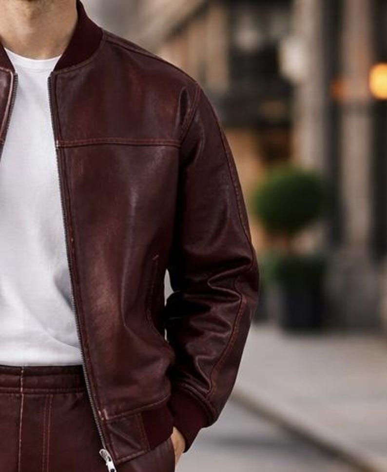 May include: A burgundy leather bomber jacket with a zip closure and ribbed cuffs. The jacket is worn over a white t-shirt. The jacket's color is a deep, rich shade of burgundy, and the leather appears to have a slight sheen.
