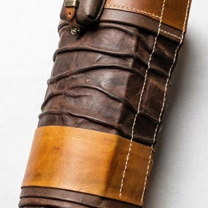 Puede incluir: Un carcaj de cuero artesanal en tonos marr&oacute;n y beige, con una peque&ntilde;a bolsa adjunta. El carcaj tiene un dise&ntilde;o texturizado y acanalado con costuras contrastantes y una ancha banda beige. Una peque&ntilde;a hebilla asegura la bolsa.