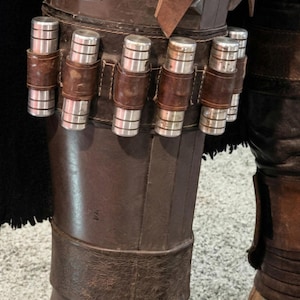 Puede incluir: Primer plano de una armadura de pierna de cuero marr&oacute;n con cartuchos cil&iacute;ndricos plateados asegurados por correas de cuero. La armadura parece desgastada y envejecida, con costuras y detalles visibles. La est&eacute;tica general sugiere un tema de ciencia ficci&oacute;n o fantas&iacute;a.