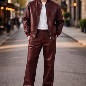 May include: A burgundy leather bomber jacket with matching pants and a white t-shirt. The jacket has a zip closure and ribbed cuffs. The pants are straight-legged. The outfit is paired with white sneakers. The model is standing on a city street.