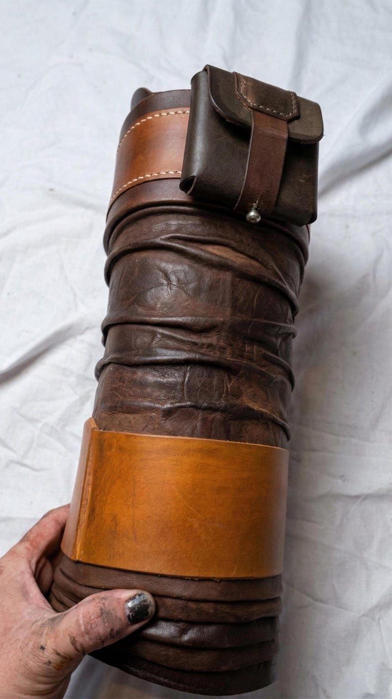 Puede incluir: Un art&iacute;culo de cuero artesanal, posiblemente un contenedor o funda, en tonos marr&oacute;n y beige. El dise&ntilde;o presenta una forma cil&iacute;ndrica con texturas en capas y una peque&ntilde;a bolsa adjunta. El art&iacute;culo se sostiene sobre un fondo blanco.