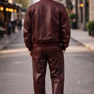 May include: A burgundy leather bomber jacket and matching pants. The jacket has a ribbed collar and cuffs, and the pants have pockets. The outfit is worn with white sneakers. The person is standing on a city street.
