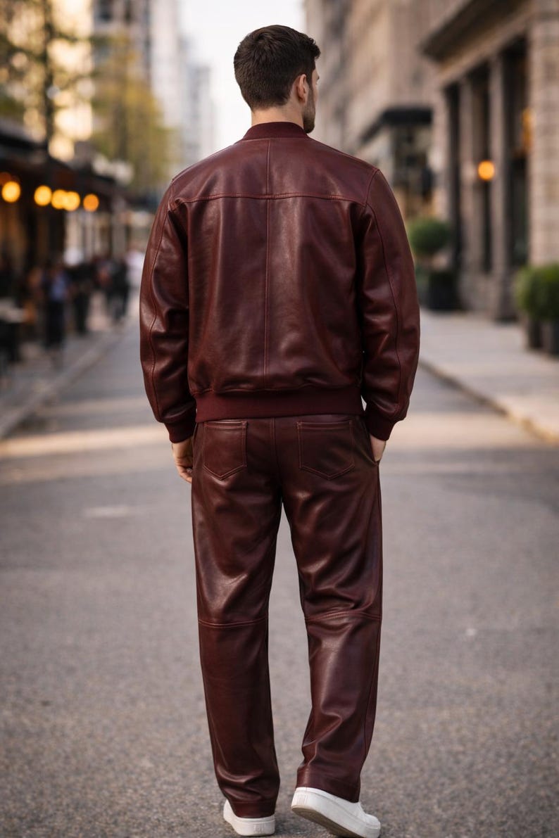 May include: A burgundy leather bomber jacket and matching pants. The jacket has a ribbed collar and cuffs, and the pants have pockets. The outfit is worn with white sneakers. The person is standing on a city street.