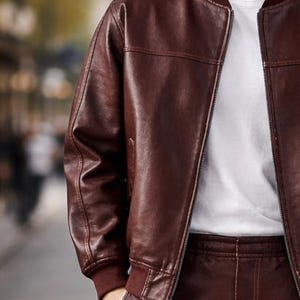 May include: A burgundy leather bomber jacket with a ribbed collar and cuffs. The jacket has a zipper closure and is worn over a white t-shirt. The leather appears smooth and has visible stitching details. The overall style is casual and modern.