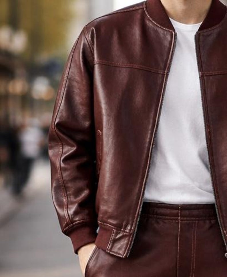 May include: A burgundy leather bomber jacket with a ribbed collar and cuffs. The jacket has a zipper closure and is worn over a white t-shirt. The leather appears smooth and has visible stitching details. The overall style is casual and modern.