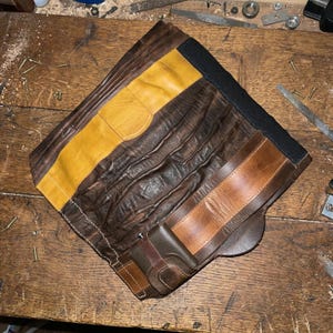 Puede incluir: Bolsa de herramientas de cuero hecha a mano con parches de cuero marr&oacute;n y amarillo. La bolsa tiene una banda el&aacute;stica negra y una peque&ntilde;a correa de cuero. El cuero tiene un aspecto envejecido.