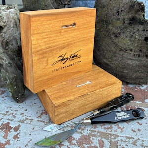 May include: Two wooden squares with a hanging hole and a signature that reads "Stacey Fabre corner of paint staceyfabre.com". Two painting tools are on the table in front of the squares.