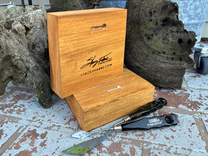 May include: Two wooden squares with a hanging hole and a signature that reads "Stacey Fabre corner of paint staceyfabre.com". Two painting tools are on the table in front of the squares.