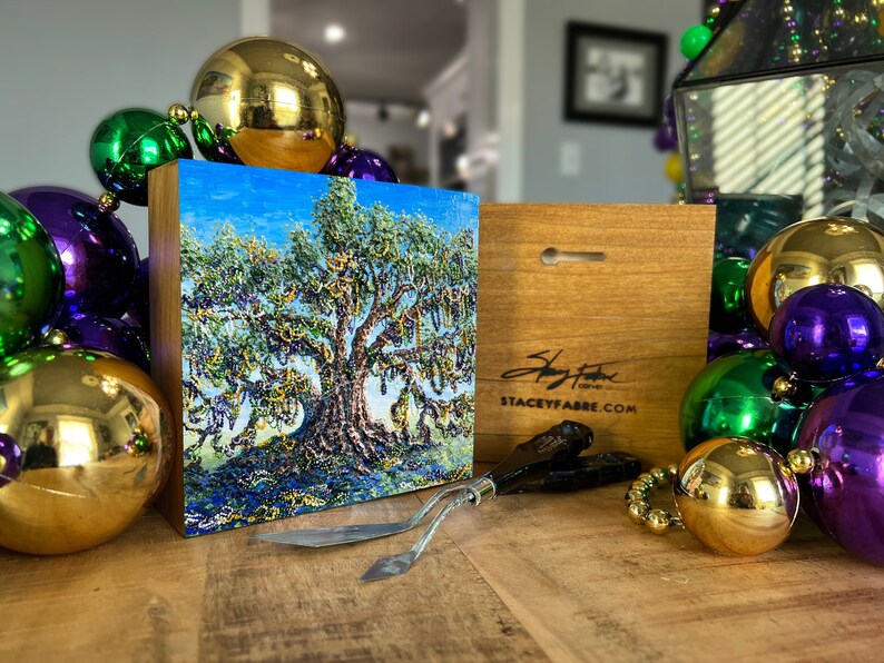 May include: A small, framed painting of a large oak tree with green leaves and blue sky. The tree is covered in purple, green, and gold beads. The painting is on a wooden base with a signature and website address on the back. The painting is surrounded by gold, green, and purple ornaments.