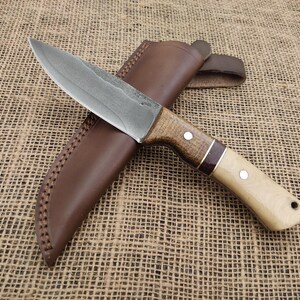 May include: A handcrafted hunting knife with a textured steel blade and a wooden handle. The handle features a light-colored wood with a dark wood accent and is paired with a brown leather sheath. The knife is designed for outdoor use.