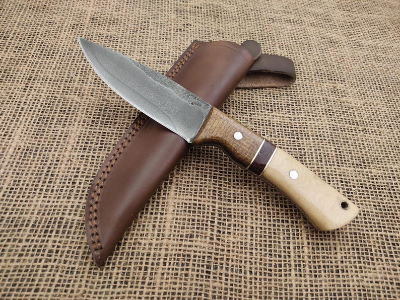 May include: A handcrafted hunting knife with a textured steel blade and a wooden handle. The handle features a light-colored wood with a dark wood accent and is paired with a brown leather sheath. The knife is designed for outdoor use.