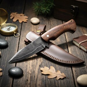 May include: A hunting knife with a dark wood handle and patterned blade sits in a brown leather sheath. A gold compass, a leather-bound journal, and decorative stones and leaves are arranged on a rustic wooden surface.