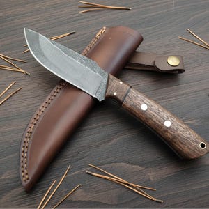 May include: A handcrafted hunting knife with a dark wooden handle and a leather sheath. The blade is silver and the handle has three white rivets. The sheath is brown and has a stitched border. Pine needles are scattered around the knife.