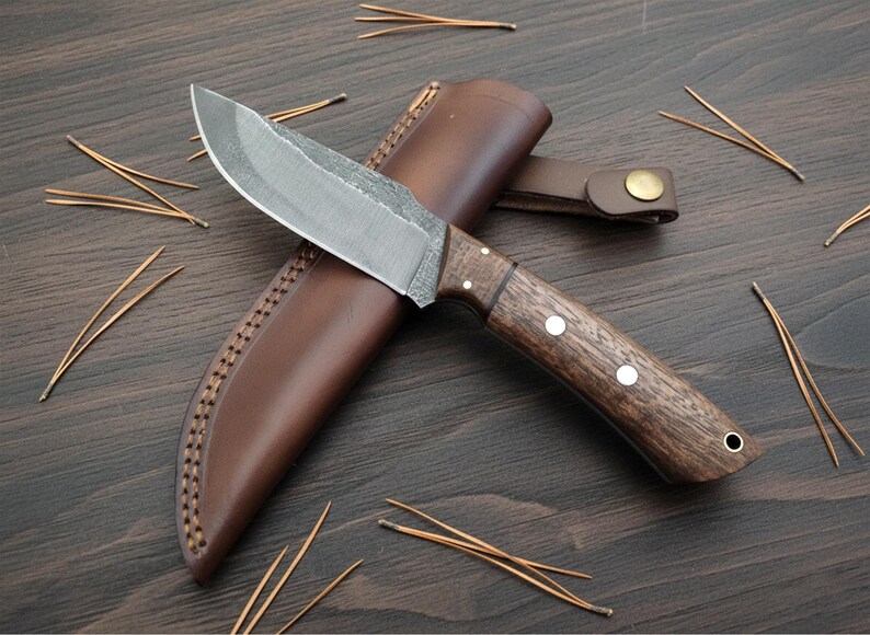 May include: A handcrafted hunting knife with a dark wooden handle and a leather sheath. The blade is silver and the handle has three white rivets. The sheath is brown and has a stitched border. Pine needles are scattered around the knife.
