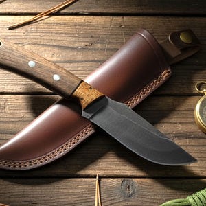 May include: A hunting knife with a dark blade and wooden handle rests on a brown leather sheath. A gold compass and green rope are also visible on a wooden surface, suggesting outdoor or survival themes.