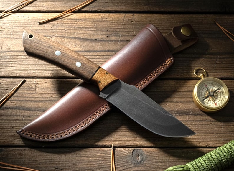 May include: A hunting knife with a dark blade and wooden handle rests on a brown leather sheath. A gold compass and green rope are also visible on a wooden surface, suggesting outdoor or survival themes.