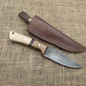 May include: A handcrafted hunting knife with a light wood handle, dark wood accents, and a metal blade. The knife is paired with a brown leather sheath. The knife and sheath are displayed on a textured, tan surface.