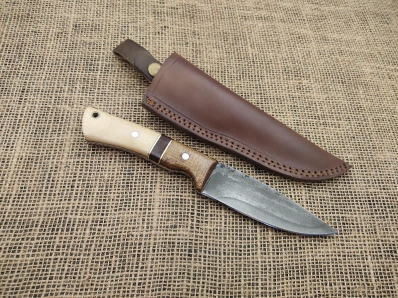 May include: A handcrafted hunting knife with a light wood handle, dark wood accents, and a metal blade. The knife is paired with a brown leather sheath. The knife and sheath are displayed on a textured, tan surface.