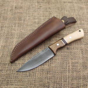 May include: A handcrafted knife with a wooden handle and a leather sheath. The blade is a dark gray color, and the handle features light wood and dark wood accents. The leather sheath is brown and has decorative stitching.