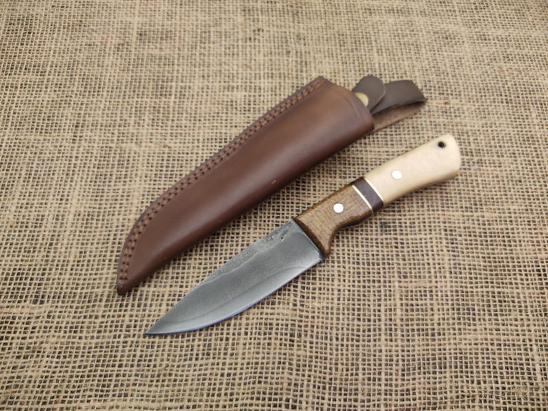 May include: A handcrafted knife with a wooden handle and a leather sheath. The blade is a dark gray color, and the handle features light wood and dark wood accents. The leather sheath is brown and has decorative stitching.