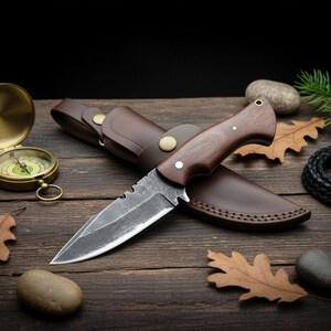 May include: A hunting knife with a dark wooden handle and a leather sheath. The blade has a serrated edge. A vintage compass and other outdoor accessories are arranged on a wooden surface.