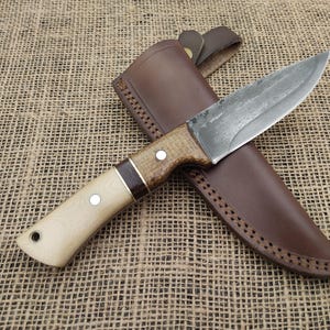 May include: A handcrafted knife with a light wood handle and a dark brown leather sheath. The blade has a rustic, textured appearance. The handle features a dark brown accent and two silver rivets. The sheath has decorative stitching.