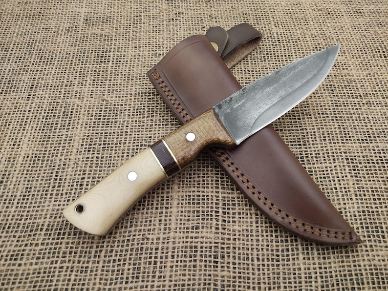 May include: A handcrafted knife with a light wood handle and a dark brown leather sheath. The blade has a rustic, textured appearance. The handle features a dark brown accent and two silver rivets. The sheath has decorative stitching.