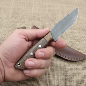 May include: A small, handcrafted knife with a wooden handle and a steel blade. The handle is a dark brown color with two silver rivets. A brown leather sheath is visible in the background.