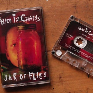 May include: A cassette tape and its case featuring the album "Jar of Flies" by Alice in Chains. The case shows a jar with a face inside. The cassette tape is clear with a red label.
