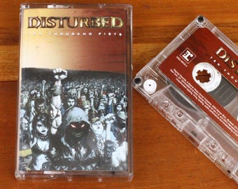 May include: A clear cassette tape with the album art for Disturbed's "Ten Thousand Fists" album. The cover art features a crowd of people with a raised fist. The cassette is on a wooden surface.
