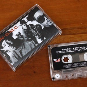 Op de afbeelding: Een transparante cassette en hoes met het album "Back From Hell" van RUN-D.M.C. De albumhoes is zwart-wit, met de gezichten van de bandleden. De cassette heeft de albumtitel en tracklist erop gedrukt. Op een houten oppervlak.