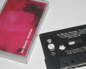 May include: A black cassette tape labeled "My Bloody Valentine / Loveless" with a clear plastic case and a red and pink cover. The cassette tape has white text and a "WB" logo. The case is open, revealing the cassette.